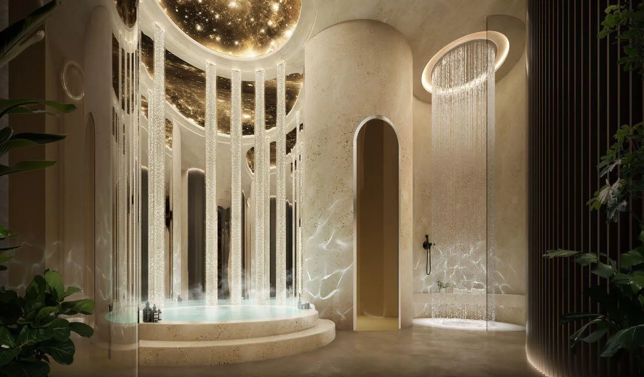 A warm-lit hall with an arched opening, a long chandelier hanging above cream stone walls and planting