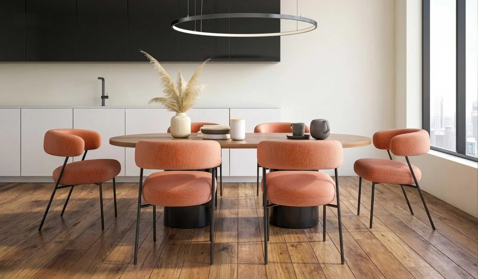 A dining table with coral upholstered chairs under a ring pendant, the kitchen units behind it
