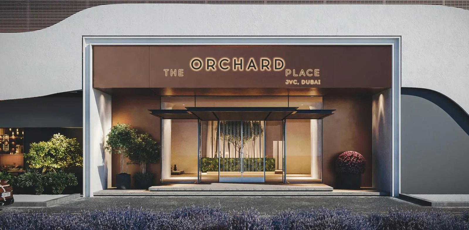 The entrance after dark, the project name lit above the doors between two beds of purple planting
