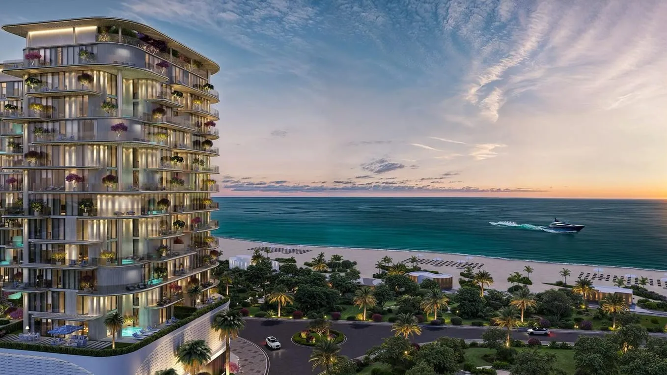 An aerial view along the shoreline at sunset, the building at the left of frame above a long stretch of sand and calm sea