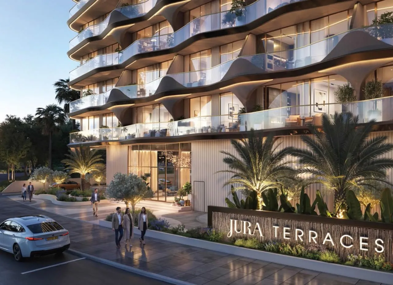 The entrance at dusk, the project's name lit on a low planted wall and people walking past palms
