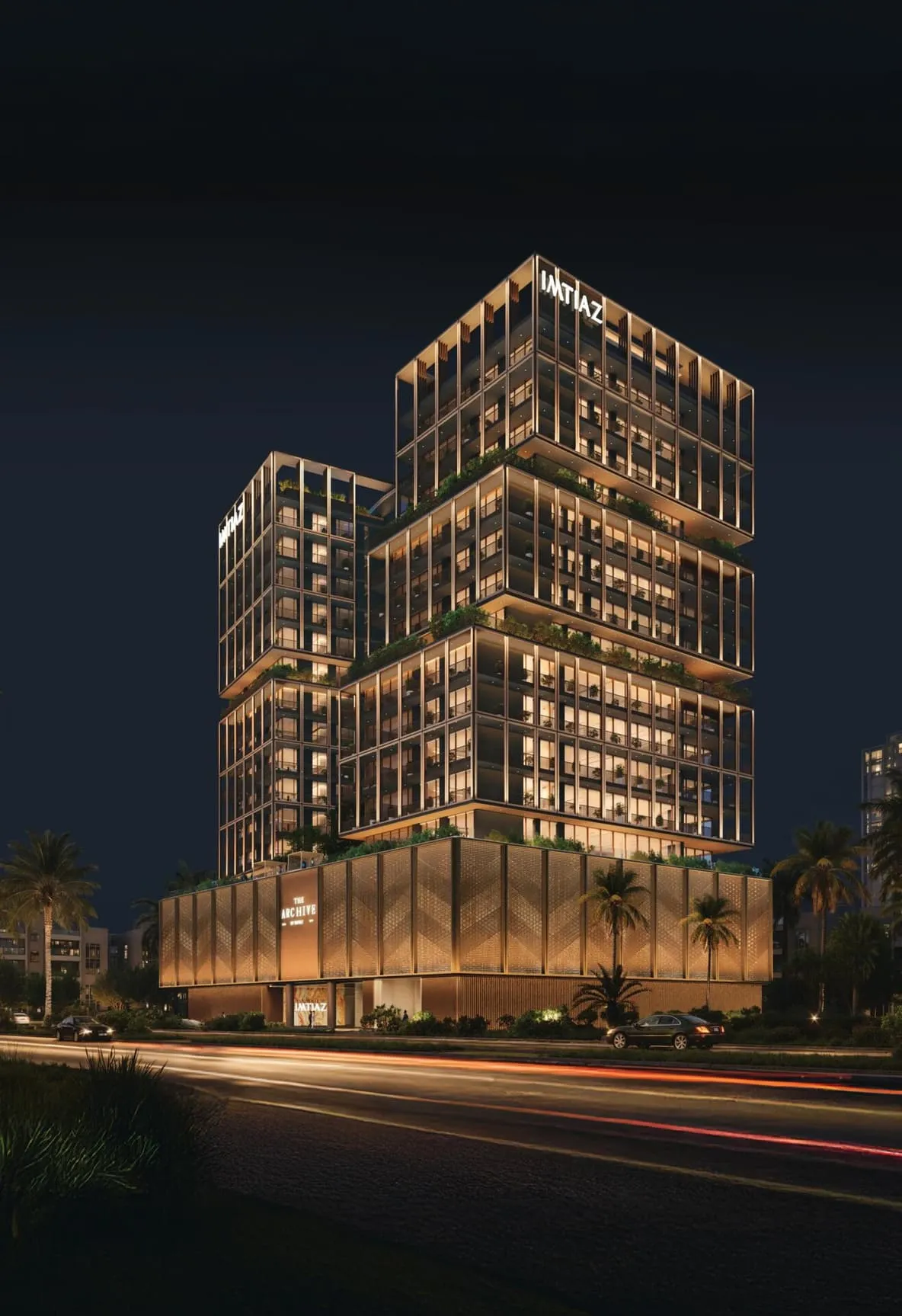 The building lit at night from the road, light trails along the carriageway and the podium screen glowing behind the palms