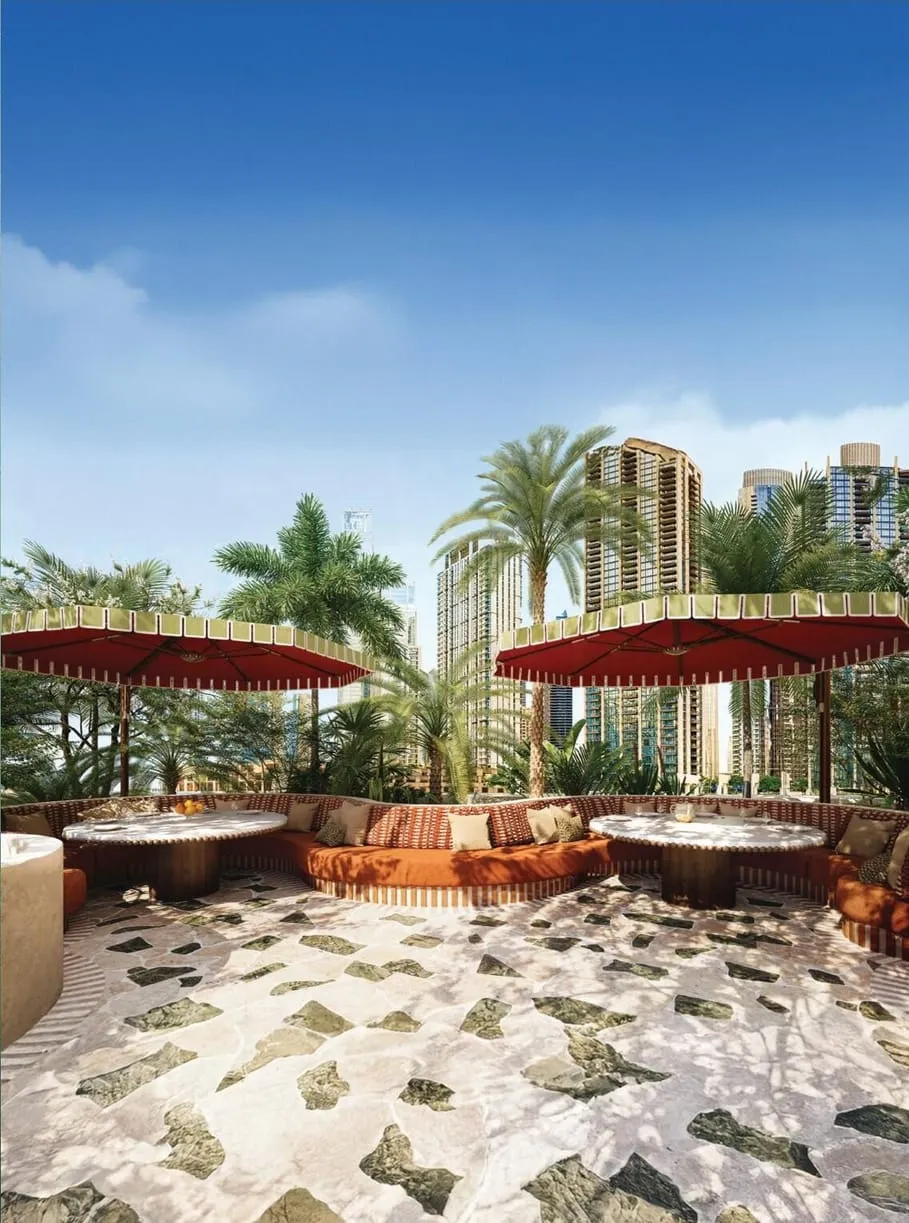 An outdoor terrace with red parasols over sunken seating, pebbles underfoot and palms against the towers
